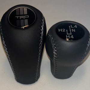 May include: Two black leather gear shift knobs with silver stitching. The larger knob features the letters "TRD". The smaller knob has gear shift markings including "H2", "L4", "N", and "H4".