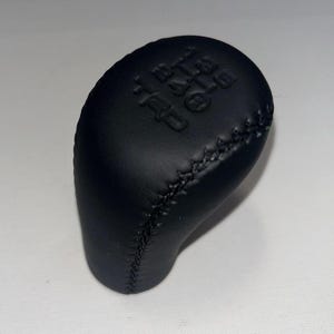 May include: Black leather gear shift knob with stitched seams. The top of the knob has the gear shift pattern and the letters "TRD" embossed on it. The knob is designed for a vehicle.
