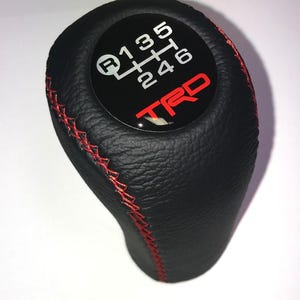 Op de afbeelding: Zwarte lederen versnellingspookknop met rode stiksels. De bovenkant heeft een zwarte cirkel met een wit schakelpatroon en een rood "TRD" logo.