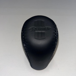 May include: Black leather gear shift knob with the word "THUNDER" and a vertical stripe design. The knob is oval-shaped and has visible stitching around the edges. The background is a plain white surface.