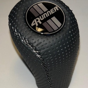 Könnte beinhalten: Schwarzer, perforierter Leder-Schaltknauf mit silbernen Nähten. Die Oberseite zeigt ein rundes Emblem mit dem Text "4Runner" in Weiß und grauen Streifen auf schwarzem Hintergrund. Für ein Fahrzeug konzipiert.