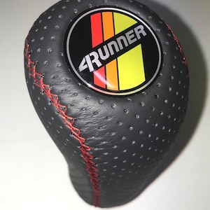 May include: A black shift knob with red stitching and a perforated surface. The knob features a circular emblem with the word "4Runner" and a design in red, orange, and yellow. Automotive accessory.