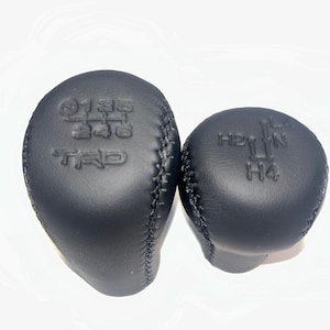 May include: Two black leather gear shift knobs. One knob has the numbers 1, 3, 5, 2, 4, 6, and the letters TRD embossed on it. The other knob has the letters H2, N, H4 embossed on it.