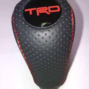 May include: A black and gray shift knob with red stitching. The top of the knob features the red letters "TRD" on a black background. The knob has a perforated texture.