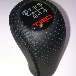 Op de afbeelding: Zwarte geperforeerde lederen versnellingspookknop met een zwart en rood TRD-logo. Het schakelpatroon wordt in het wit op een zwarte achtergrond weergegeven. Ontworpen voor een handgeschakelde versnellingsbak.