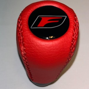 May include: Red leather shift knob with a black emblem featuring a white outline and a red "F" logo. The knob has a stitched seam and a black base. Automotive accessory.