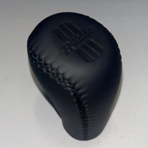 Op de afbeelding: Een zwarte, met leer omwikkelde versnellingspookknop met een gestikte naad. De bovenkant van de knop heeft de tekst "Rumbles" in reliëf boven horizontale lijnen. De knop is ontworpen voor de versnellingspook van een voertuig.
