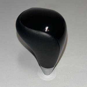 Op de afbeelding: Een glanzende zwarte versnellingspookknop met een zilveren basis. De knop heeft een gladde, afgeronde bovenkant en een voorgevormde vorm voor comfortabele bediening. De zilveren basis zorgt voor een contrasterend accent op de zwarte afwerking.