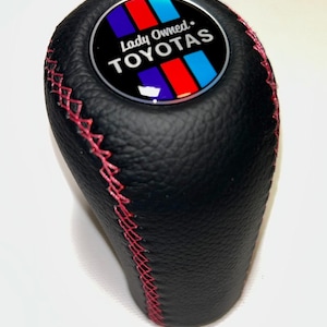 Peut inclure: Pommeau de levier de vitesse en cuir noir avec surpiqûres rouges. Le dessus présente un emblème circulaire avec le texte "Lady Owned. TOYOTAS" et un motif de rayures bleues, rouges et blanches. Le pommeau est pour un véhicule.