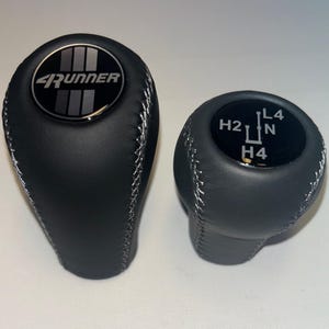 Op de afbeelding: Twee zwarte lederen versnellingspookknoppen. De grotere knop heeft het woord "4Runner" in zilveren letters op een zwart cirkelvormig embleem. De kleinere knop heeft versnellingsmarkeringen: H2, L4, N en H4. Beide knoppen hebben witte stiksels.