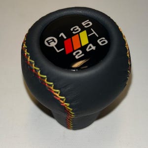 Op de afbeelding: Een zwarte leren versnellingspookknop met een kleurrijk schakelpatroon. Het schakelpatroon is zwart met witte cijfers en een rode, oranje en gele streep. De knop heeft gele en rode stiksels.