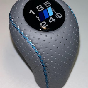 May include: Grey perforated leather gear stick knob with blue stitching. The top has a black circle with the gear shift pattern in white, and a blue and white logo. Designed for a manual transmission.