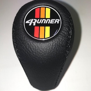 Könnte beinhalten: Schwarzer Lederschaltknauf mit einer Naht. Der Knauf hat ein rundes Emblem mit dem weißen Text "4Runner" und vertikalen Streifen in Rot, Orange und Gelb.
