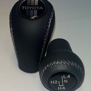 May include: Two black leather gear shift knobs. The larger knob has the Toyota logo on top. The smaller knob has gear shift markings: H2, L4, N, and H4. Both knobs have silver stitching.