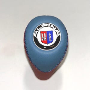 May include: A gear shift knob featuring a blue leather top section with blue stitching and a central circular emblem. The emblem displays the text ALPINA in white letters around a shield divided into red and blue halves with white details.