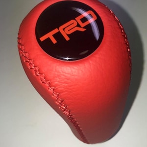 May include: Red leather gear shift knob with the letters "TRD" in orange on a black circular background. The knob has a stitched seam and a smooth, rounded shape.