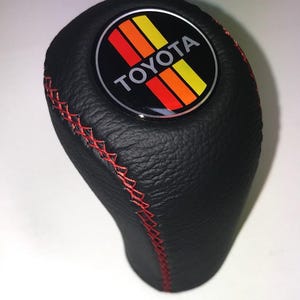 May include: Black leather gear stick knob with red stitching. It has a circular emblem with the Toyota logo and a retro design in red, orange, and yellow. The stitching provides a contrasting detail to the black leather.