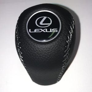 Könnte beinhalten: Schwarzer Lexus-Schaltknauf aus Leder mit silbernen Nähten. Das Lexus-Logo befindet sich mittig auf der Oberseite des Knaufs, eingefasst in einem silbernen Kreis. Ersatzteil für ein Fahrzeug.