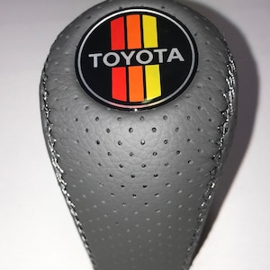 Peut inclure: Un pommeau de levier de vitesses en cuir gris perforé avec des coutures argentées. Il présente un emblème circulaire avec le logo Toyota et des rayures verticales rouges, orange et jaunes. Le texte "TOYOTA" est en blanc.