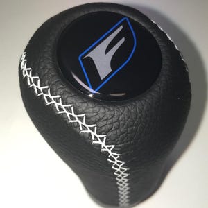 May include: Black leather shift knob with white stitching. The top features a black emblem with a silver "F" logo outlined in blue. The knob has a textured surface and a rounded shape.