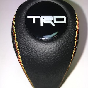 May include: Black shift knob with the letters "TRD" in white on a glossy black surface. The knob is made of textured black leather and has yellow and red stitching around the edges.