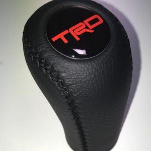 Op de afbeelding: Zwarte lederen versnellingspookknop met rood "TRD" logo op een zwarte cirkelvormige achtergrond. De knop heeft een gestikte naad en een getextureerd oppervlak. Auto-accessoire.