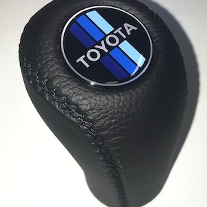 May include: Black leather gear shift knob with a circular emblem. The emblem features the TOYOTA logo in white text, with blue and light blue stripes on a black background. The knob has a textured surface.