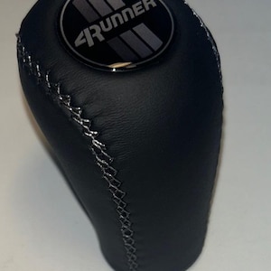 May include: Black leather shift knob with silver stitching. The top of the knob features a black circular emblem with the word "4Runner" in silver lettering and a silver stripe design.