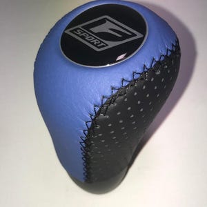 May include: A blue and black leather shift knob with the word "SPORT" and the letter "F" in silver on a black circular emblem. The shift knob has a stitched design.