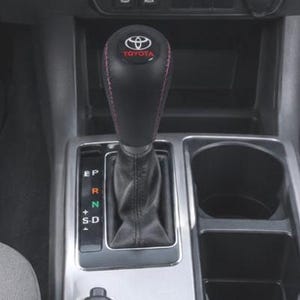 Peut inclure: Un pommeau de levier de vitesse en cuir noir avec des surpiqûres rouges et le logo Toyota sur le dessus. Le sélecteur de vitesse affiche les lettres P, R, N, D et S en couleurs, intégré dans une console argentée à côté de deux porte-gobelets.