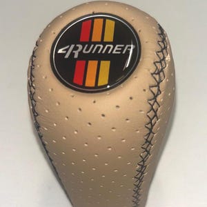 Peut inclure: Un pommeau de levier de vitesse en cuir beige perforé avec un emblème circulaire sur le dessus. L'emblème affiche le texte 4RUNNER en lettres blanches, entouré de bandes verticales rouges, orange et jaunes sur fond noir. Des coutures foncées.