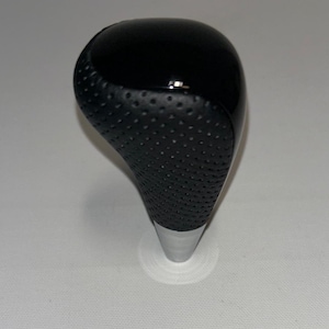 Op de afbeelding: Een zwarte versnellingspookknop met een geperforeerde grip en een glanzende bovenkant. De basis is zilver. Dit auto-onderdeel is ontworpen voor de transmissie van een voertuig.