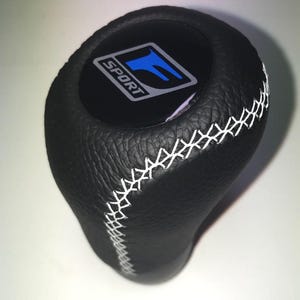 May include: Black leather shift knob with white stitching. The top features a black emblem with the word "SPORT" and a blue "F" logo. The shift knob is designed for a vehicle's gear shift.
