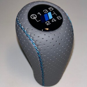 May include: Gray perforated leather shift knob with blue stitching. The top features a black circle with the gear shift pattern in white, blue, and black. The shift pattern includes R, 1, 2, 3, 4, 5, and 6.
