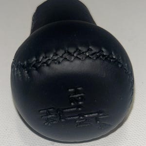 May include: A black gear shift knob featuring a spherical top with a textured cross-stitch pattern and an embossed gear pattern on the surface showing H, 2, 4, and L positions. The knob has a smooth matte finish and a rounded, ergonomic shape.