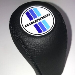 May include: Black leather shift knob with a stitched seam. The knob features a white circular emblem with the text "4RUNNER" and blue stripes. The shift knob is designed for a vehicle.