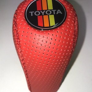 May include: A red perforated leather shift knob with the TOYOTA logo. The logo is a black circle with the word TOYOTA in white and vertical stripes of orange, yellow, and red. The shift knob is a vibrant red color.