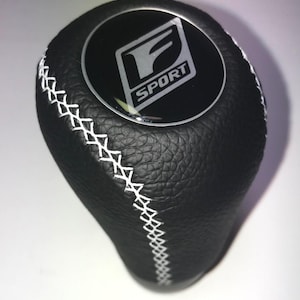 May include: Black leather shift knob with white stitching. The knob has a circular emblem with the letters "F SPORT" in silver. The shift knob is designed for a vehicle's interior.