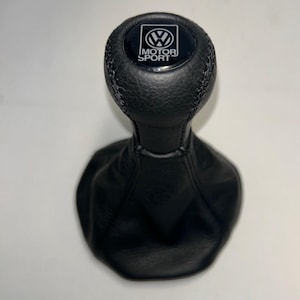 May include: A black leather gear shift knob with a matching black leather boot. The top of the knob features a circular black emblem with the Volkswagen logo and the text MOTOR SPORT printed in white lettering, surrounded by a smooth black border.