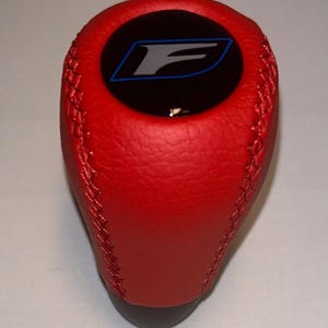 May include: Red leather shift knob with a black top featuring a blue and white "F" logo. The knob has a stitched design and a black base. Automotive accessory.