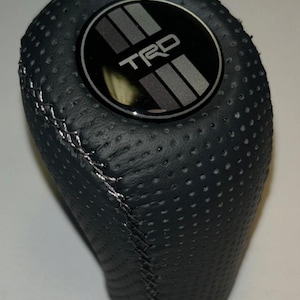 May include: Black, perforated leather shift knob with silver stitching. The top features a circular emblem with the letters "TRD" and silver stripes. The knob is designed for a vehicle's gear shift and has a textured surface for grip.