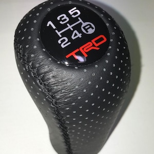 May include: Black leather gear stick knob with a perforated pattern. The top has a black circle displaying white gear numbers and the letters "R" and "F". The word "TRD" is in red.