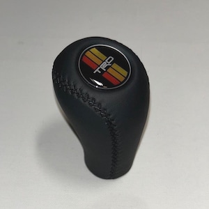 May include: A black gear shift knob featuring a circular emblem on top with the text TRD in white letters, set against a background of three horizontal stripes in yellow, orange, and red. The knob has visible black stitching along the side.