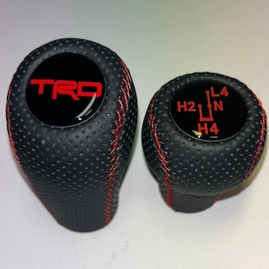 Könnte beinhalten: Zwei schwarze Schaltknäufe aus perforiertem Leder mit roten Nähten. Der größere Knauf hat eine glänzend schwarze Oberseite mit dem roten Schriftzug TRD. Der kleinere Knauf hat eine glänzend schwarze Oberseite mit einem roten Schaltschema H2, H4, L4, N.