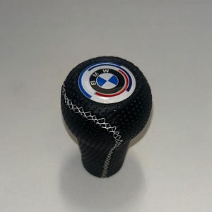 May include: A black perforated leather gear shift knob featuring a white stitched seam running diagonally across the surface. The top of the knob displays a circular BMW logo with blue, white, and red accents on a white background.
