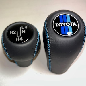 May include: Two black leather gear shift knobs with blue stitching. One knob features the Toyota logo and blue stripes. The other has white gear shift markings.