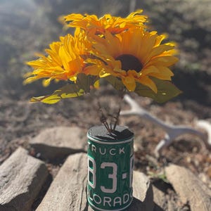 May include: A floral arrangement featuring bright yellow sunflowers in a vase made from a green Colorado license plate. The vase displays the number 83-1 in white lettering. The sunflowers' petals are a vivid yellow, with dark brown centers.