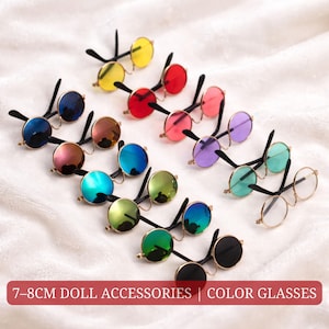Puede incluir: Una colección de gafas de muñeca coloridas con lentes redondas en varios tonos, incluyendo amarillo, rojo, rosa, morado y azul. Las gafas tienen monturas doradas y patillas negras. Texto en la imagen: 7-8cm Doll Accessories | Color Glasses.