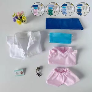 Puede incluir: Un conjunto de batas y accesorios médicos para muñecas en miniatura. Incluye una camisa blanca abotonada, uniformes rosas, un gorro quirúrgico azul, una mascarilla y un pin de estetoscopio. También se muestran varios conjuntos de uniformes de diferentes colores.