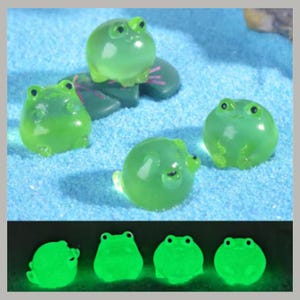 以下が含まれることがあります： 黒い目を持つ4つの半透明の緑色のカエルの置物が、水色の表面に表示されています。1匹のカエルが緑色のスイレンの葉の上に座っています。画像の下部には、緑色に光るカエルが表示されています。